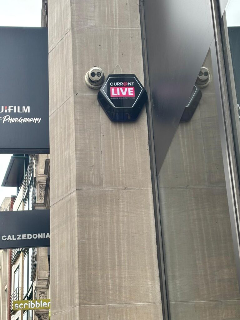 A Currainlive security camera and branded monitoring plaque mounted on a commercial storefront to deter crime and provide external surveillance.