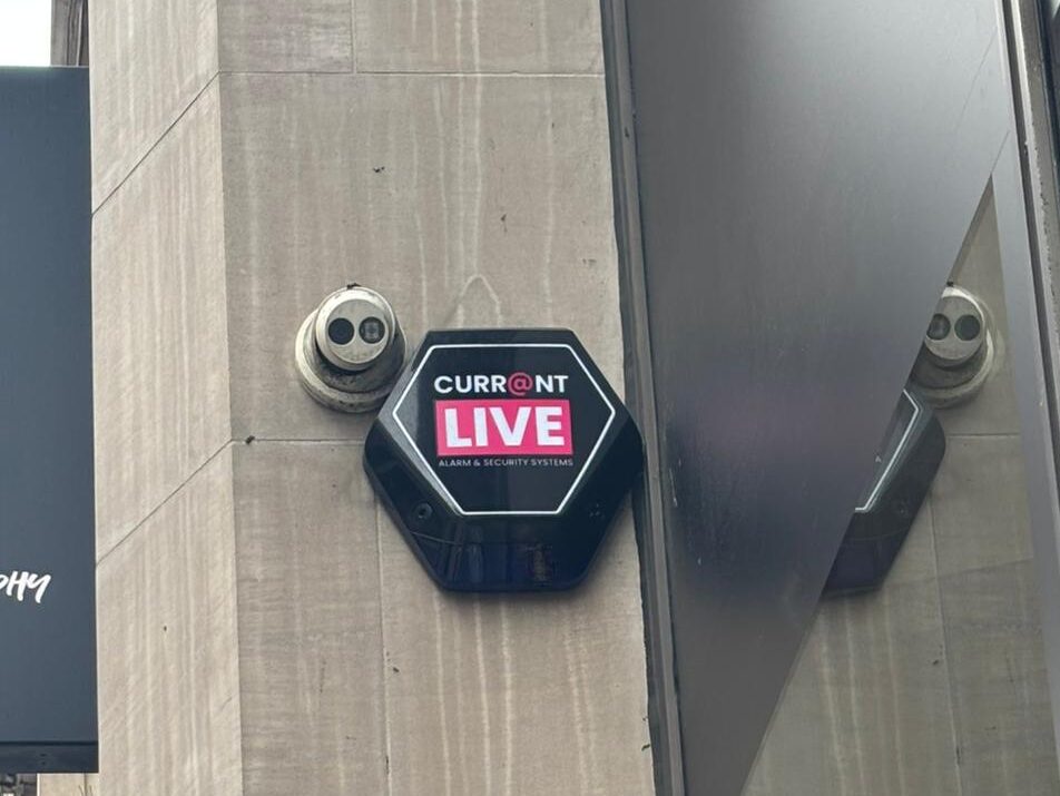 A Currainlive security camera and branded monitoring plaque mounted on a commercial storefront to deter crime and provide external surveillance.