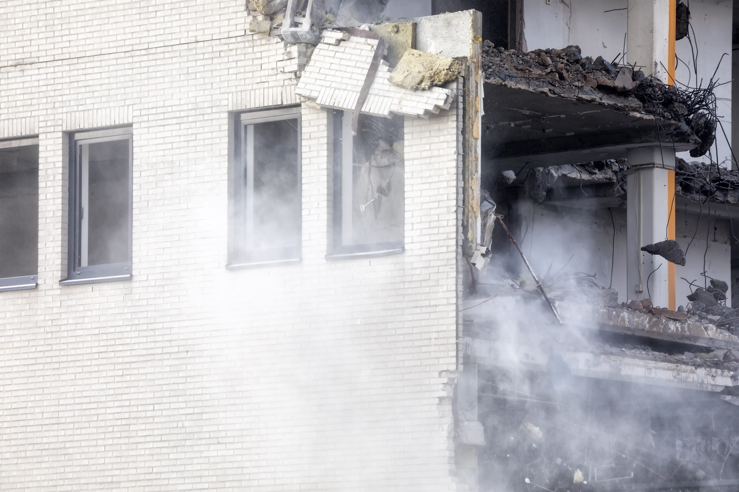 Partially destroyed building with a white brick wall, broken floors, and smoke rising from rubble at the base.